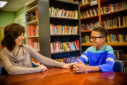 A woman and a boy are seated at a table, surrounded by bookshelves filled with various books.