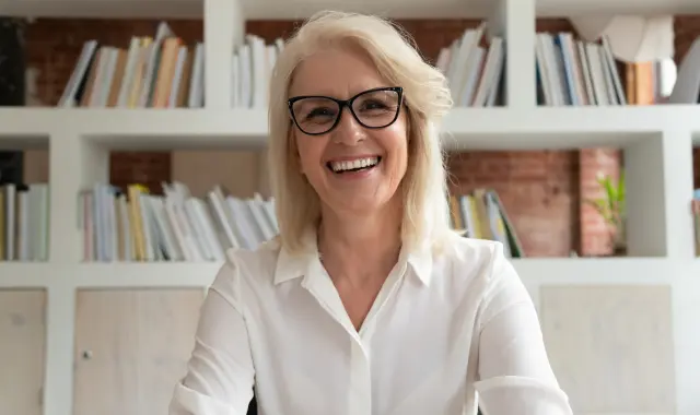 A woman wearing glasses sits at a desk, surrounded by bookshelves filled with various books.