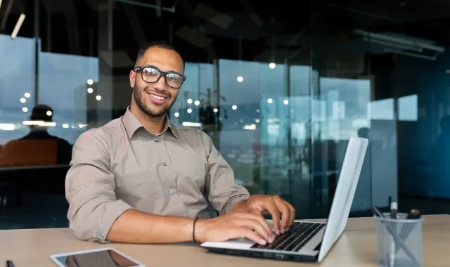 A smiling man wearing glasses sits at a desk, focused on his laptop, creating a warm and inviting workspace atmosphere.