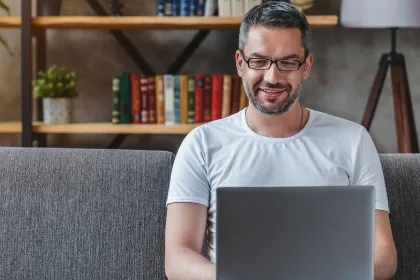 A man wearing glasses sits comfortably on a couch, focused on his laptop in a cozy living room setting.