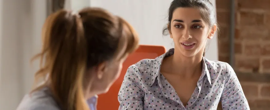 Two women engaged in conversation at a table in a professional office setting, discussing work-related matters.