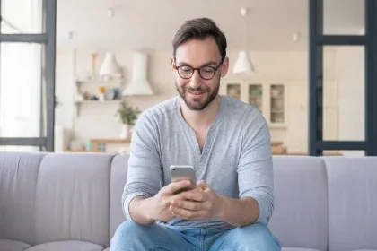 A man wearing glasses sits comfortably on a couch, focused on his phone as he engages with its screen.