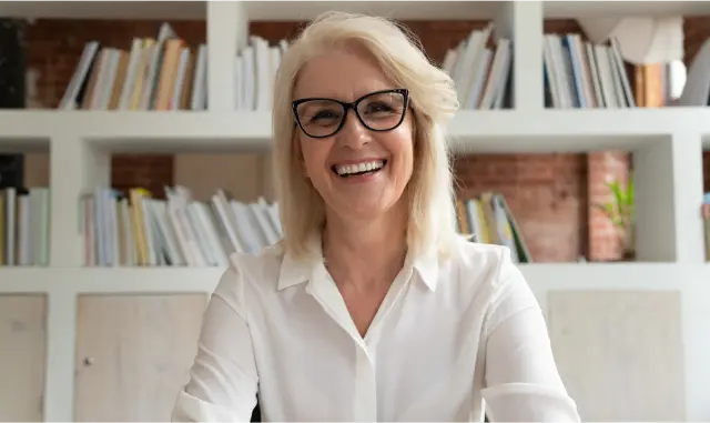 A cheerful woman with glasses looks directly at the camera, showcasing a friendly and inviting smile.