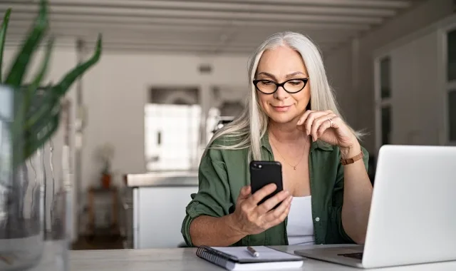 A woman with glasses sits at a table, engaged with her laptop and cell phone, demonstrating a productive workspace.