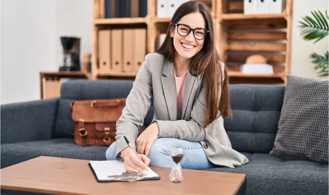 A woman wearing glasses sits on a couch, focused on writing in her notepad, surrounded by a cozy living room setting.